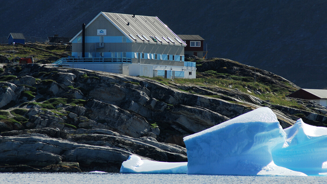 Hotel Disko Bay på klippekant med isbjerg i forgrunden og bjerg i baggrunden.