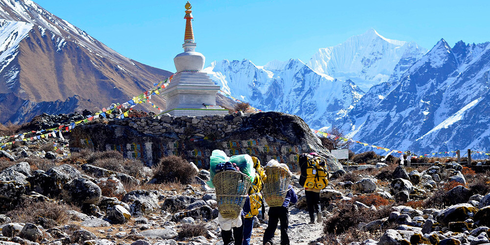 Mennesker, der vandrer mod Langtang gennem store moræne af sten og grus, med klar blå himmel og snefylde bjerge i baggrunden.