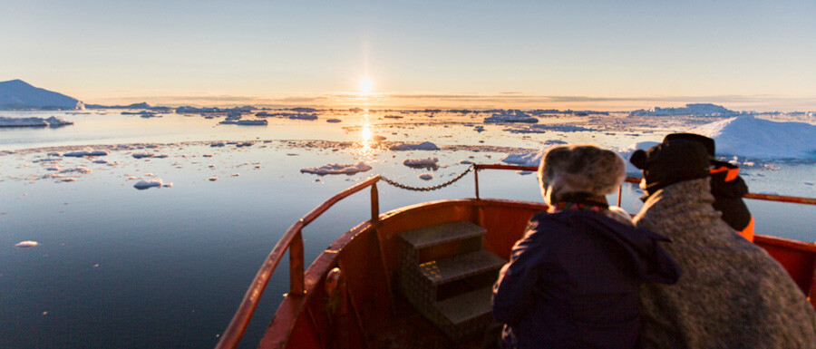 To personer står ved spidsen af et skib og ser solnedgang over isfjorden