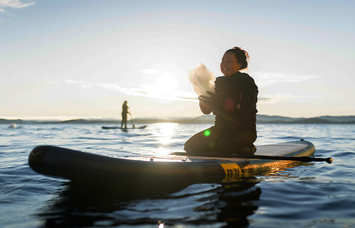 A smiling woman sitting on a SUP board in the water, with blue sky and sun shining from the background.