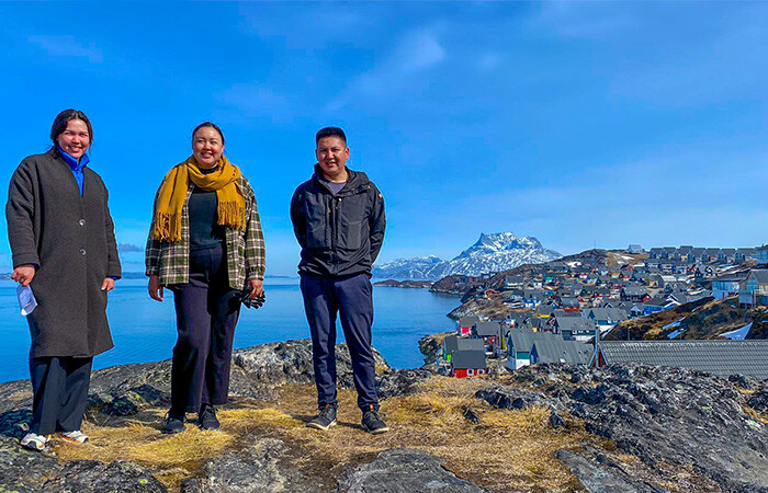 3 smiling people standing on a small hill, with a beautiful view of the city and it's colorful houses in the background. Bright blue sky and ocean in the backgorund.