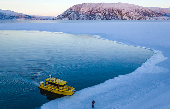 Yellow boat holding still by the ice in the ocean, with a  person standing on the ice next to the boat. Beautiful view of the blue ocean and ice, and colorful mountain in the background because of sunset.